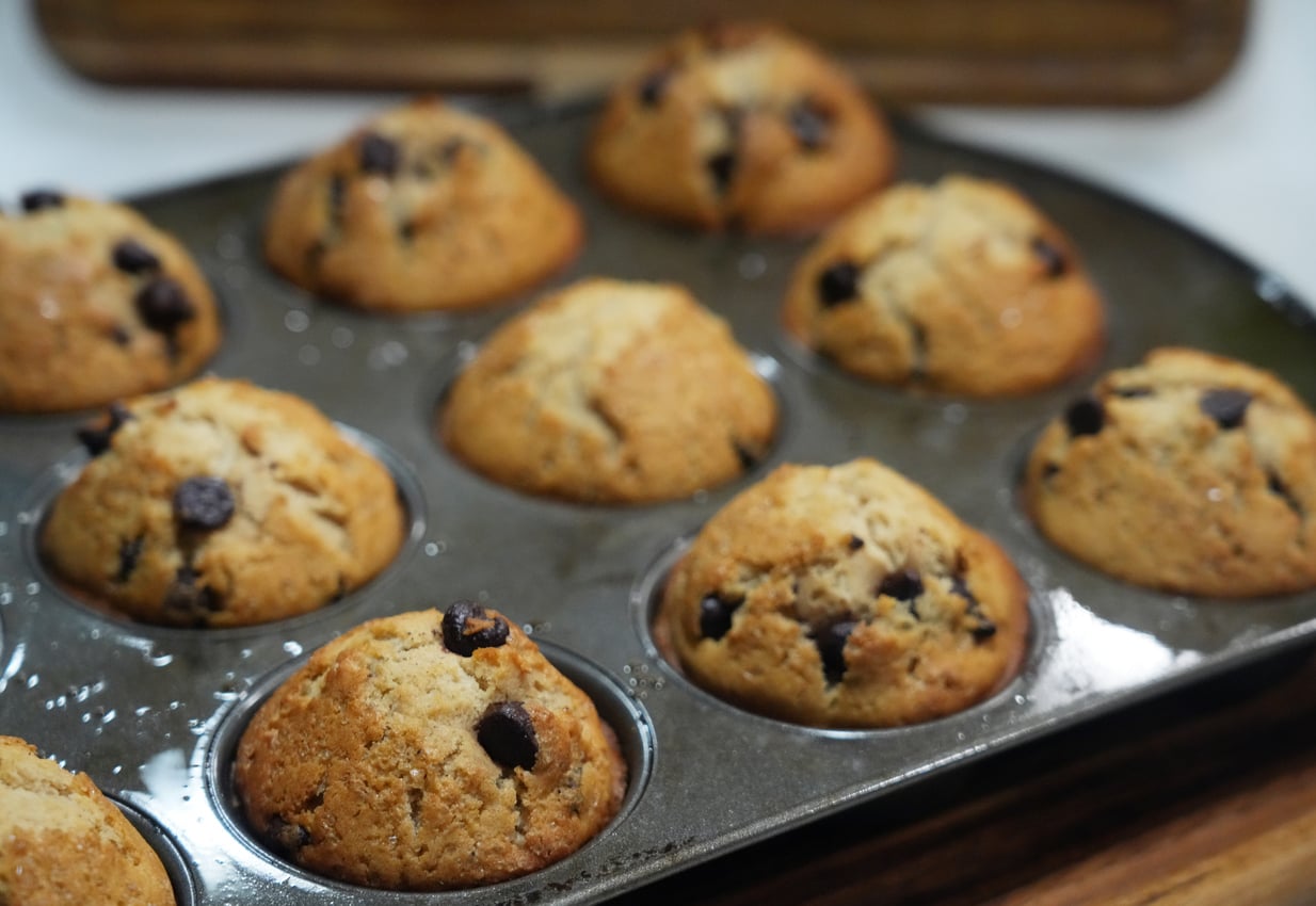 A close-up of a muffin tin filled with freshly baked chocolate chip muffins, golden brown and slightly domed, resting on a wooden surface.