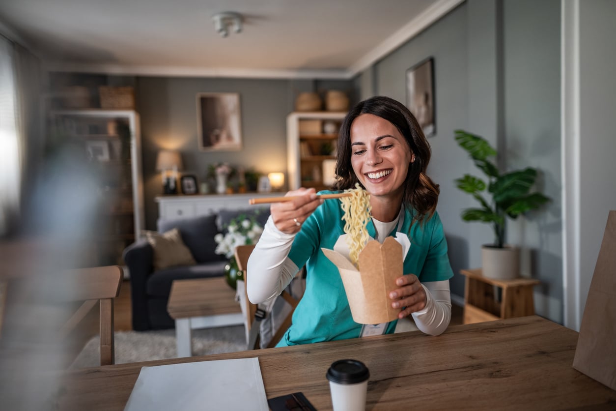 A woman smiling and eating noodles with chopsticks from a takeout box—a popular choice among foods to order—while sitting at a table in a cozy, well-lit living room.