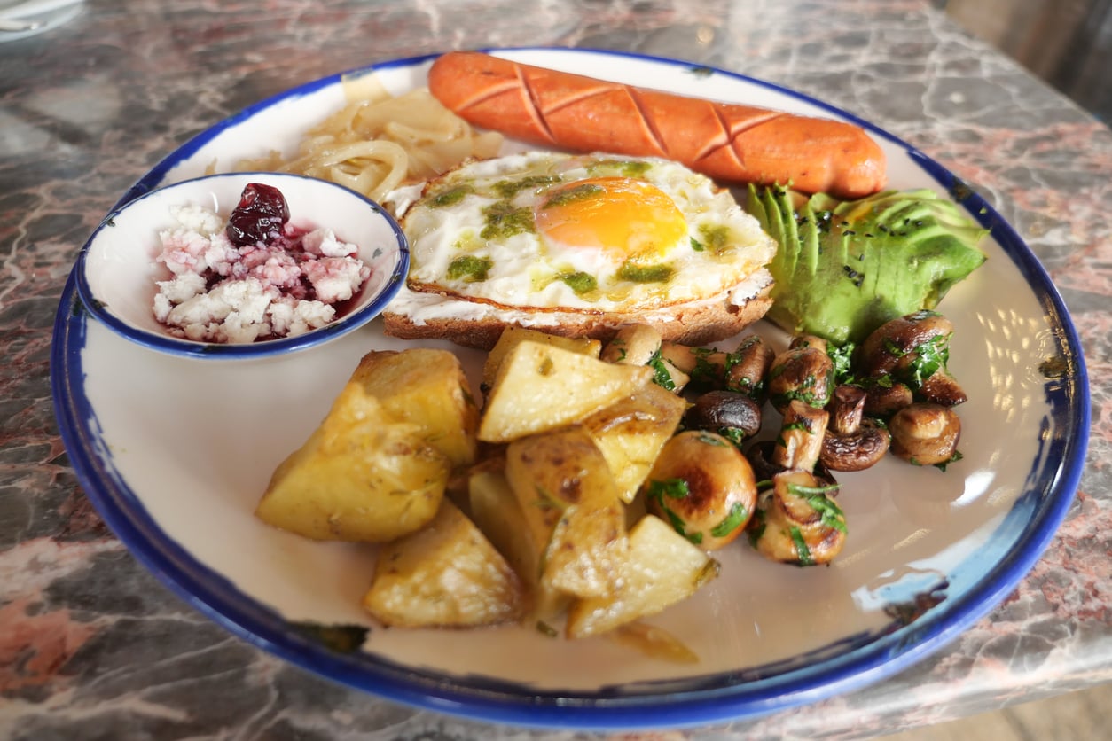 A plate with a sunny-side-up egg on toast, grilled sausage, avocado slices, roasted potatoes, sautéed mushrooms, onions, and a small bowl of crumbled cheese with berry sauce, on a marble table.