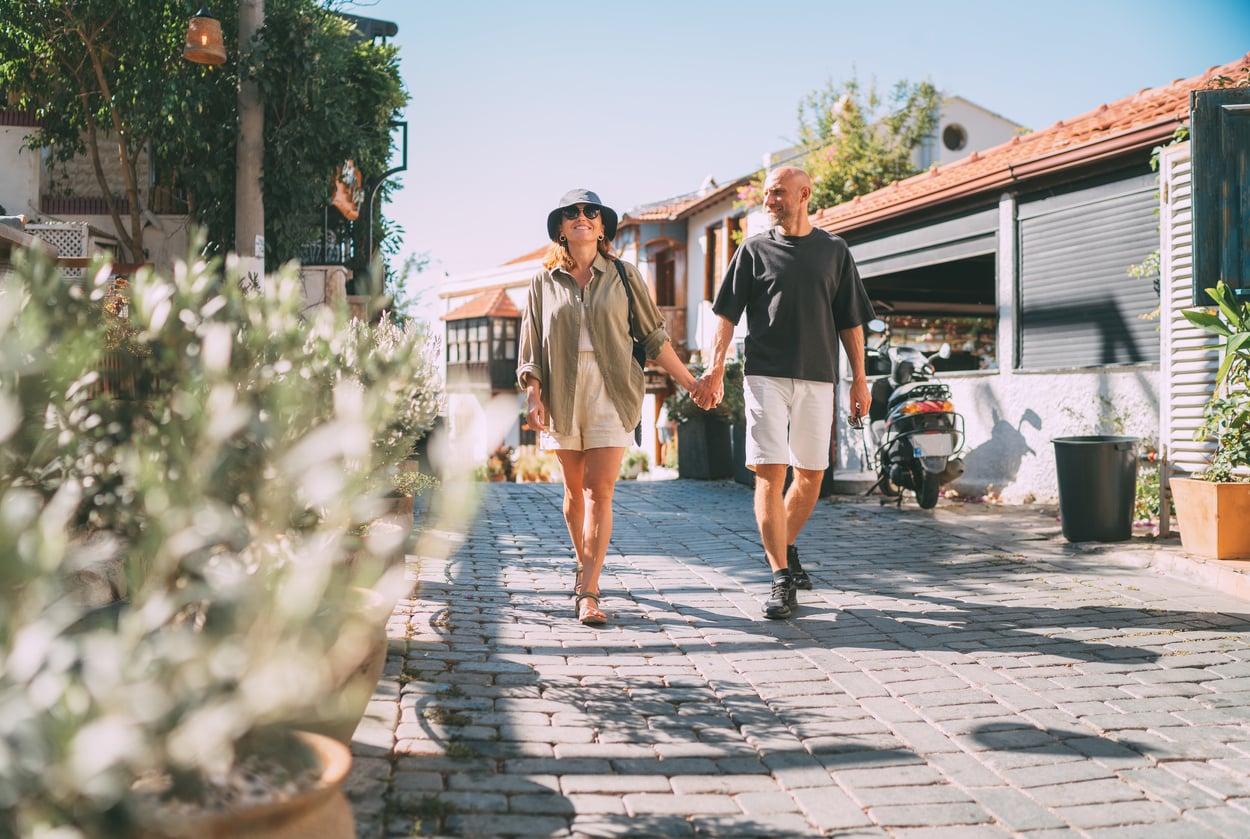 A couple walks hand-in-hand down a sunny, cobblestone street lined with plants and houses, enjoying a relaxed day outdoors—a perfect example of cherished vacation experiences.