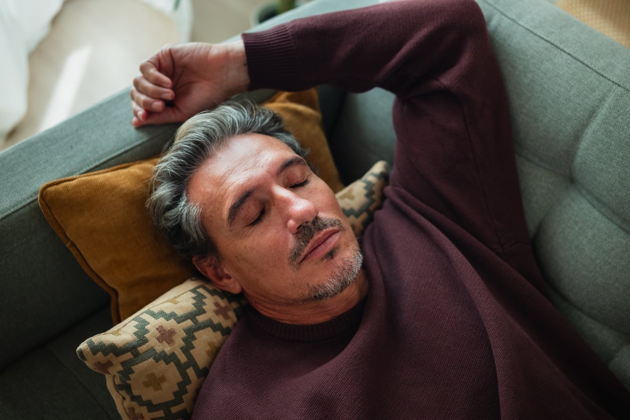 A middle-aged man with gray hair is lying on a couch with his eyes closed, resting his head on a pillow while wearing a maroon sweater.