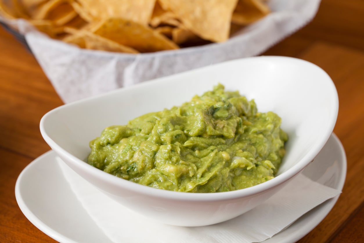 A white bowl of guacamole sits on a napkin with tortilla chips in a basket blurred in the background on a wooden table.