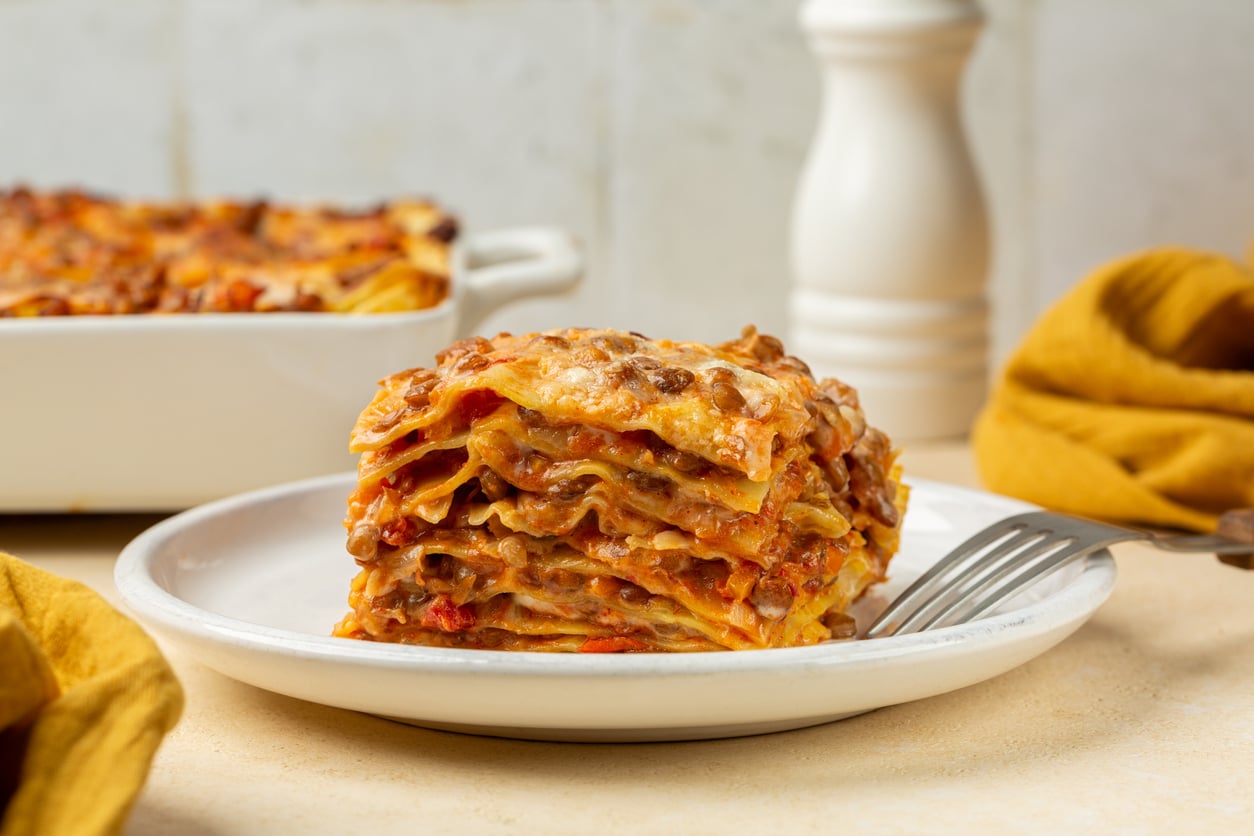 A slice of lasagna with layers of pasta, meat sauce, and melted cheese sits on a white plate with a fork. A casserole dish and yellow napkin are in the background.
