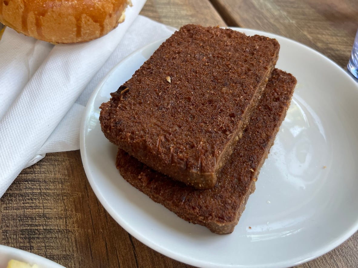 Two rectangular slices of dark brown bread are stacked on a white plate, sitting on a wooden table next to a napkin and part of a pastry.