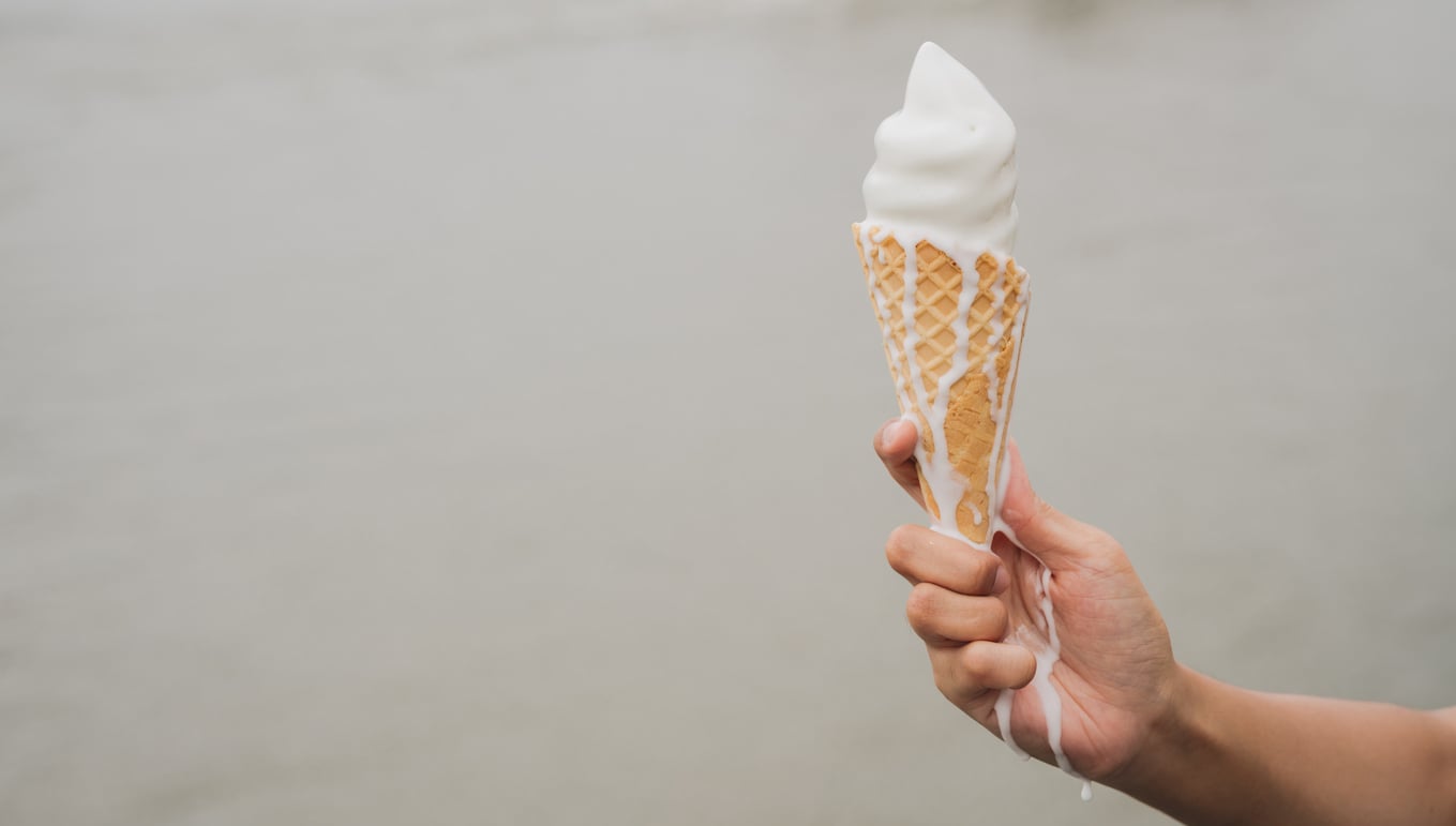 A hand holds a melting vanilla soft serve ice cream cone, with ice cream dripping down the cone against a blurred, neutral background.