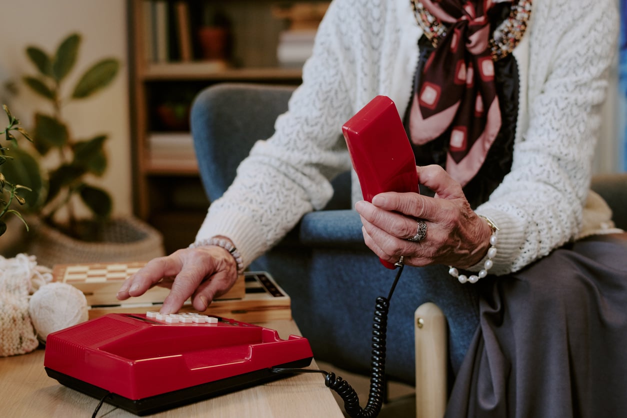 An elderly person in a sweater and scarf sits on a chair, holding a red corded telephone and dialing a number, with a chessboard and plant in the background.