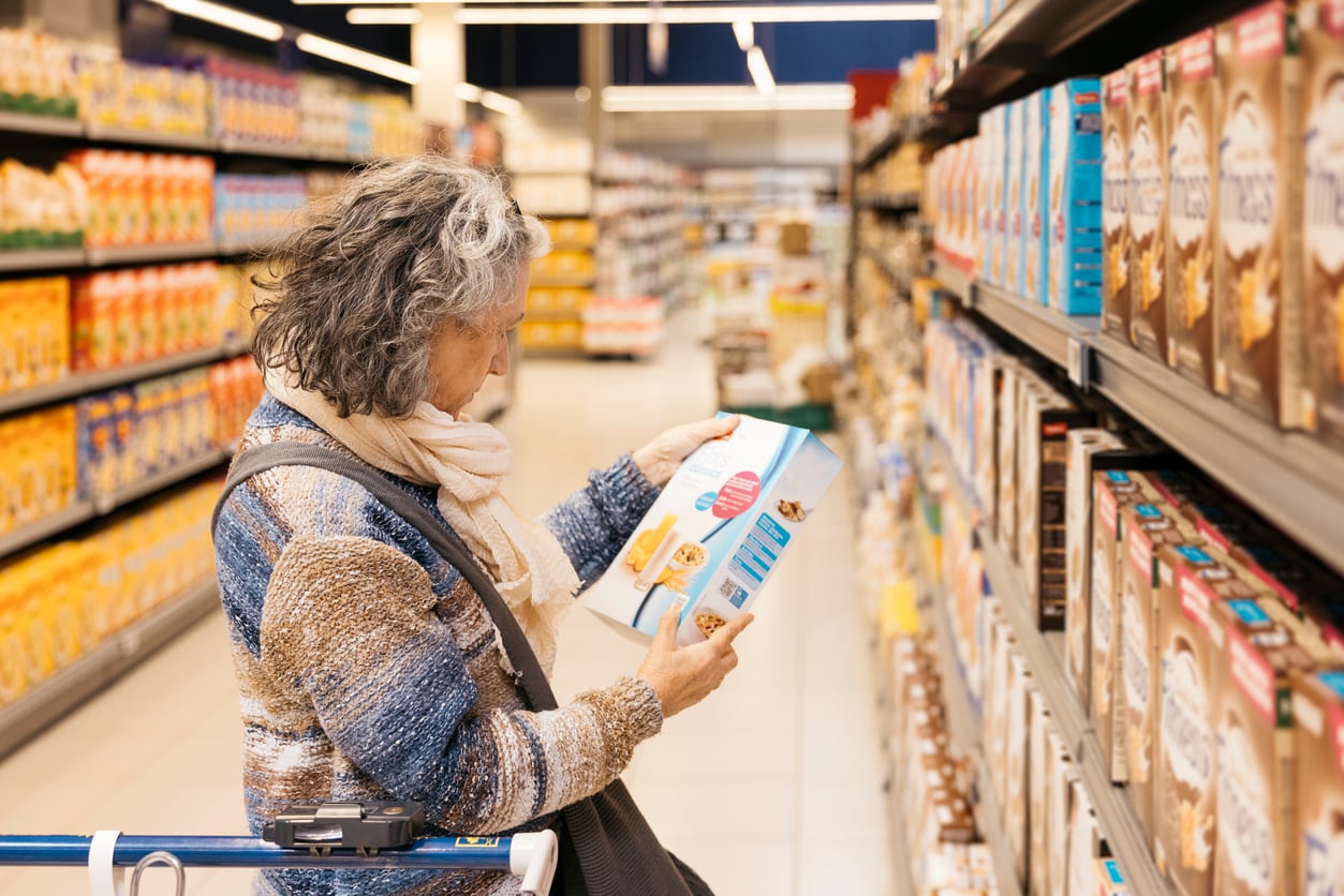 An older woman with gray hair and a scarf examines a cereal box in a grocery aisle filled with various cereals.