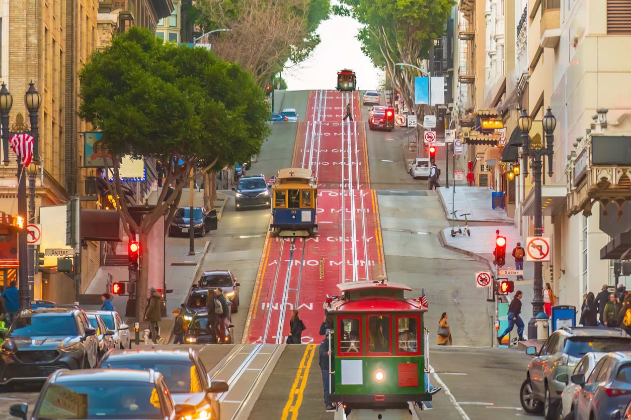 San Francisco street with steep hill, cable cars traveling up and down, traffic lights, parked cars, and buildings on both sides. Green trees and pedestrians visible along sidewalks.