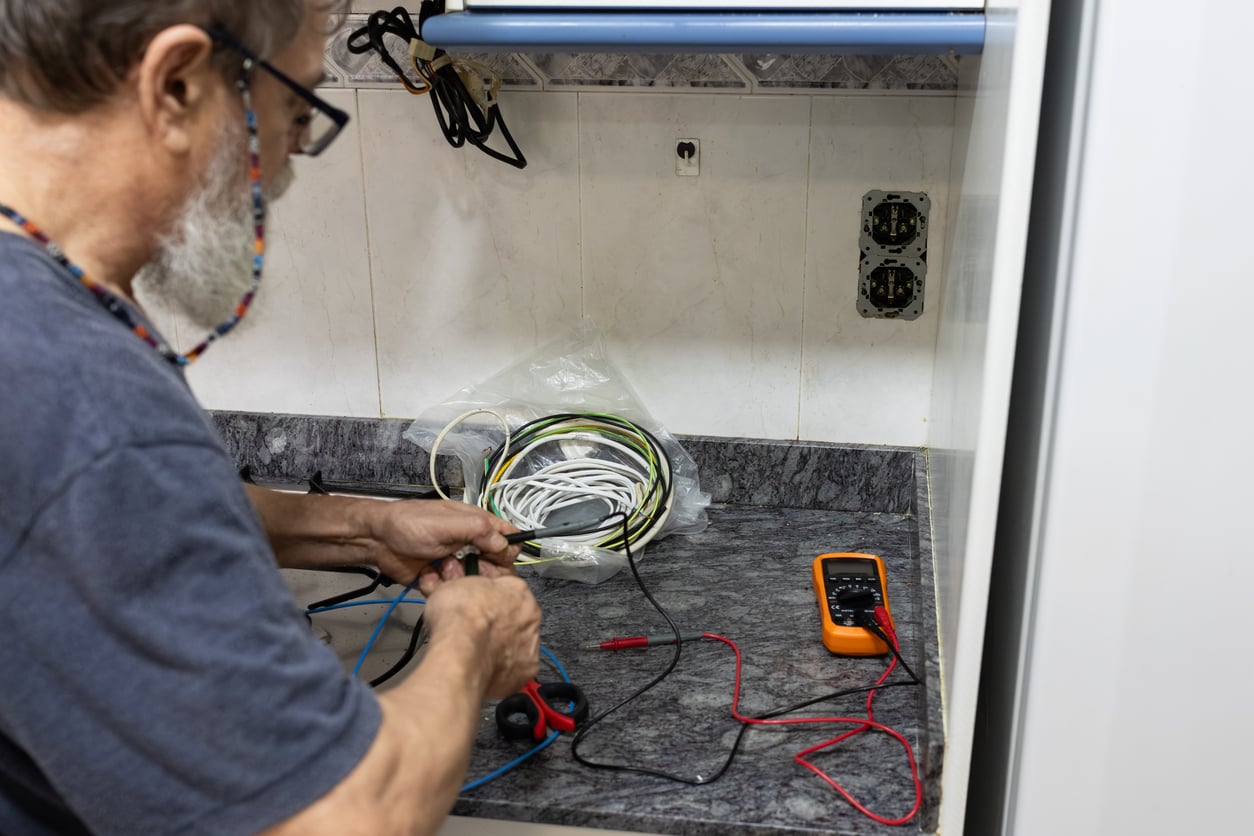 A person working with electrical wires on a kitchen counter, using tools and a digital multimeter, with open outlets on the wall nearby.