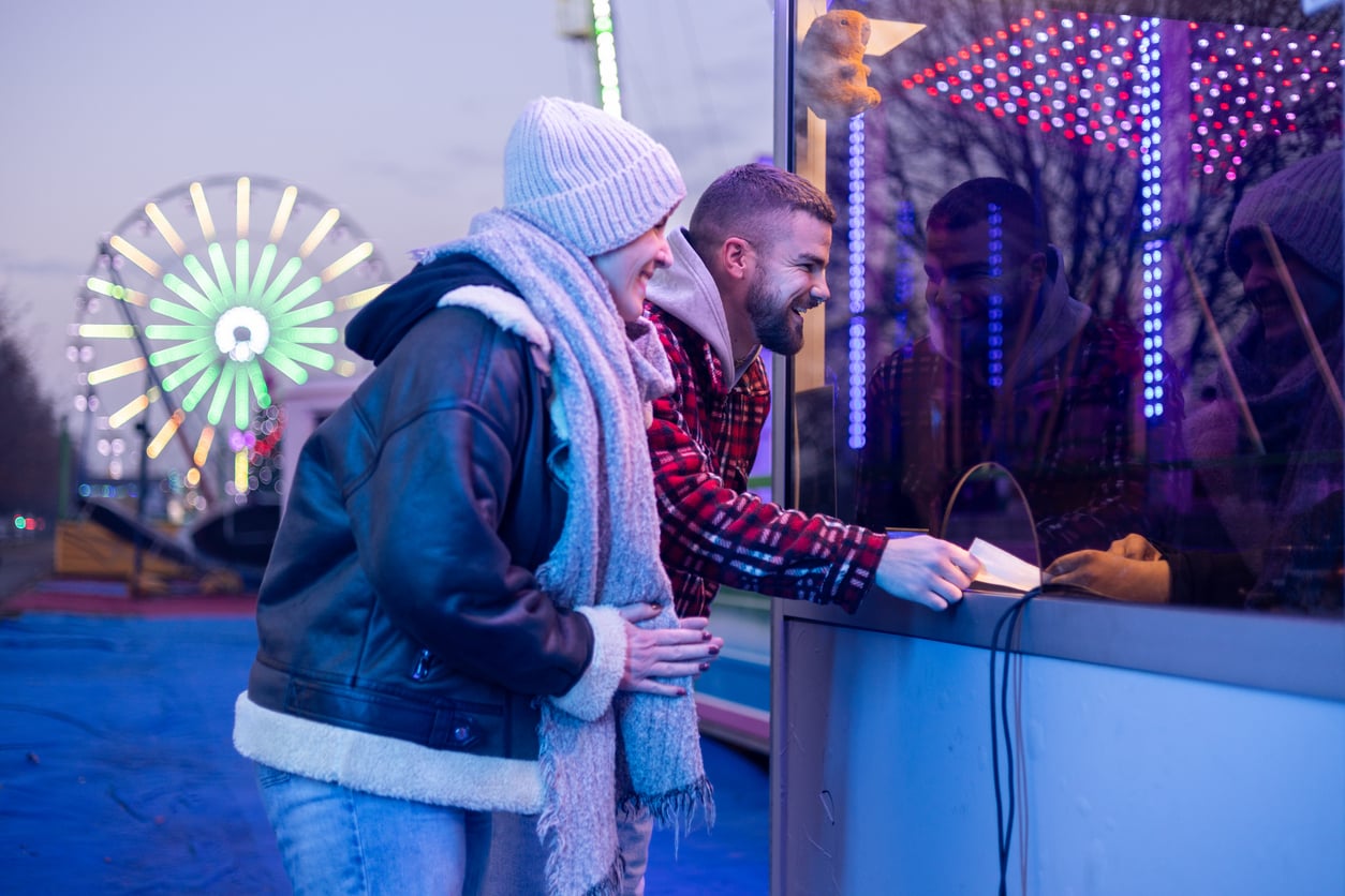Two people in winter clothing smile as they buy tickets at a booth at a fairground, with colorful lights and a Ferris wheel glowing in the background.