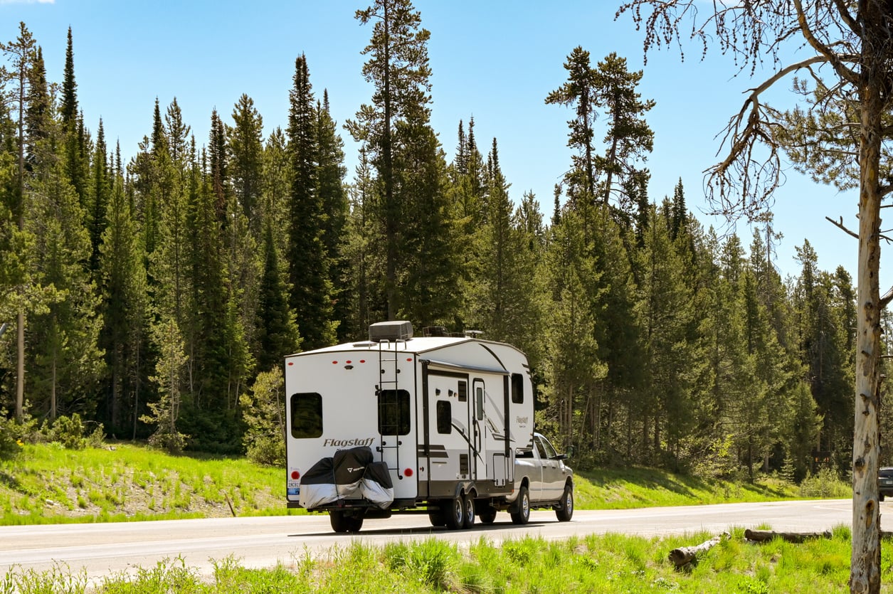 A white pickup truck towing a large white camper travels down a road bordered by tall pine trees and greenery under a clear blue sky.