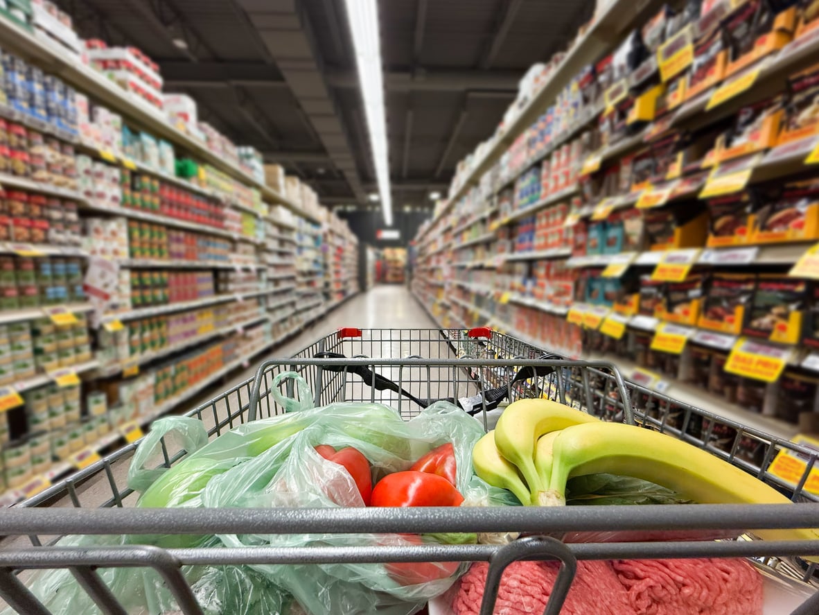 A shopping cart with bananas, tomatoes, and grocery bags inside sits in the middle of a brightly lit supermarket aisle lined with shelves of various food products, perfectly capturing everyday grocery habits.