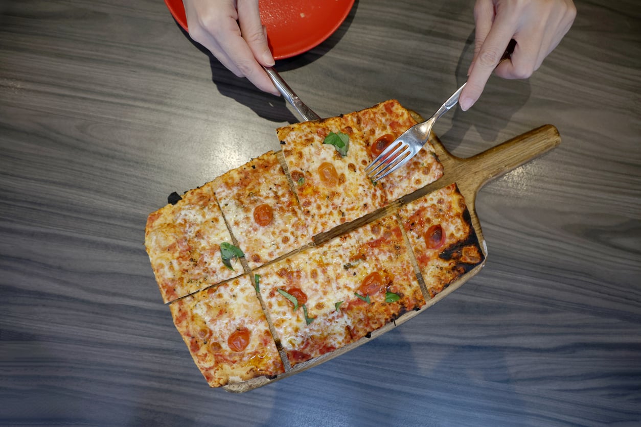 A person cuts a rectangular pizza with a fork and knife on a wooden board. The pizza has cheese, cherry tomatoes, and basil. A red plate sits nearby on a gray table.