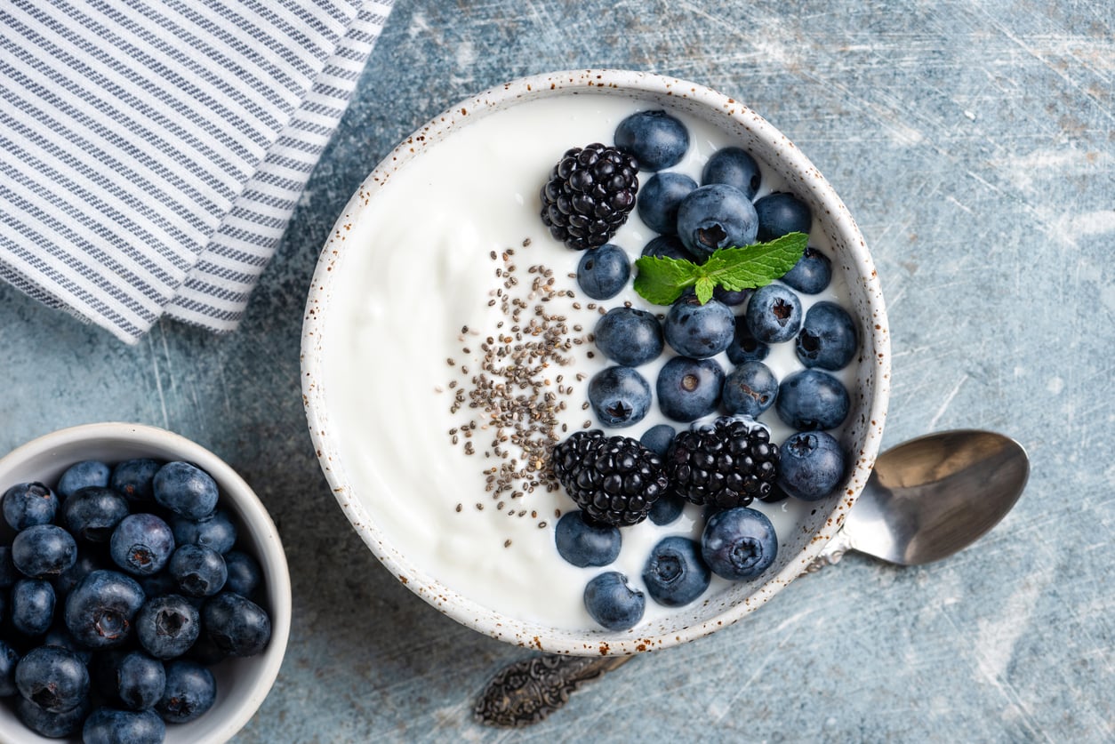 A bowl of yogurt topped with blueberries, blackberries, chia seeds, and a mint leaf—a perfect example of everyday foods that used to be considered fancy—sits next to a small bowl of blueberries, a spoon, and a striped napkin on a blue textured surface.