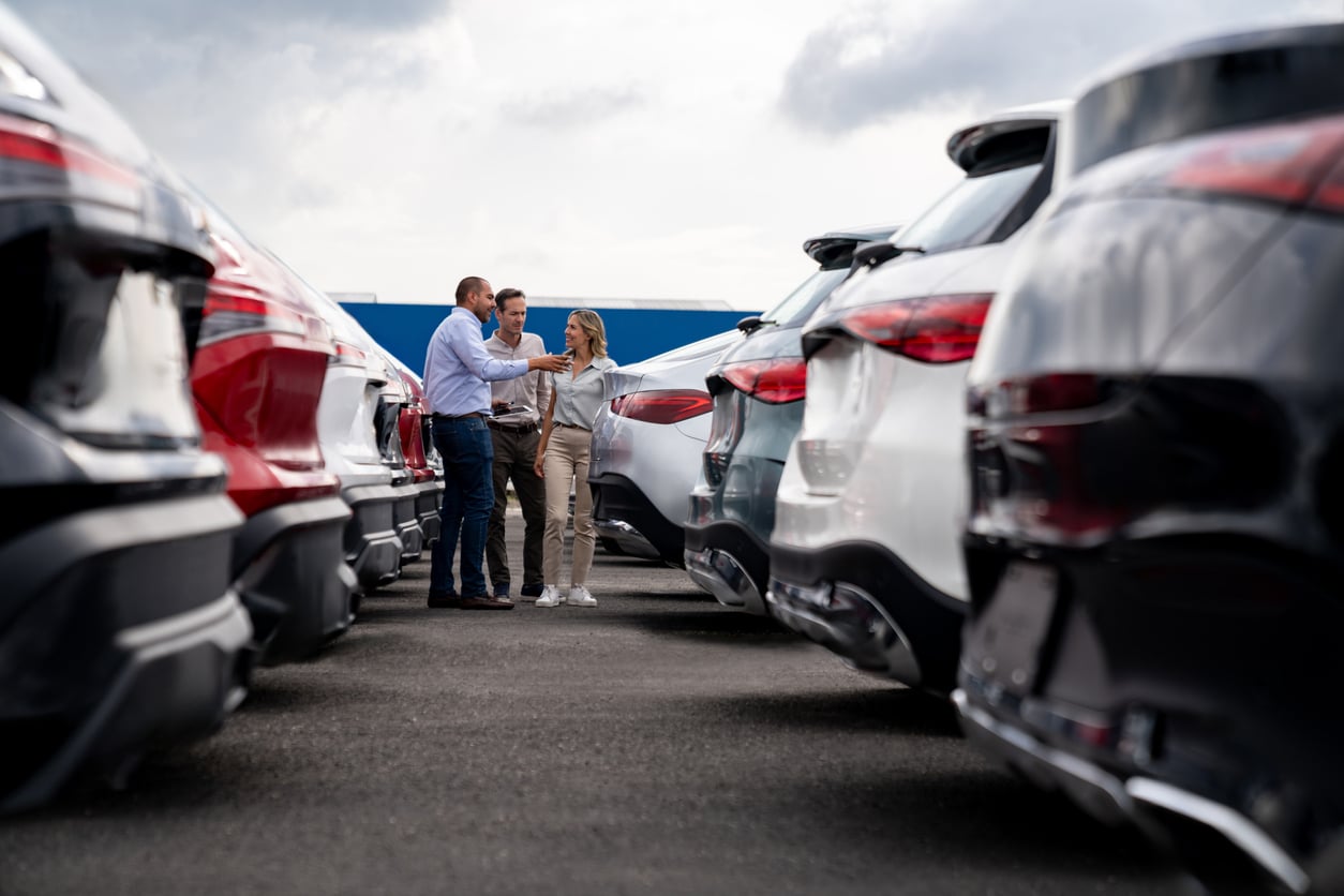 A car salesperson talks to a couple among rows of parked cars at a dealership lot on a cloudy day.