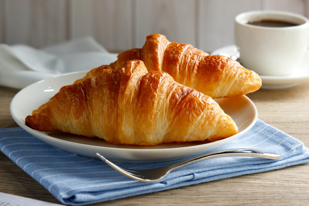 Two golden croissants on a white plate, with a fork and a blue napkin, sit on a wooden table—an inviting display of everyday foods that used to be considered fancy. A cup of coffee and a white napkin are in the background.
