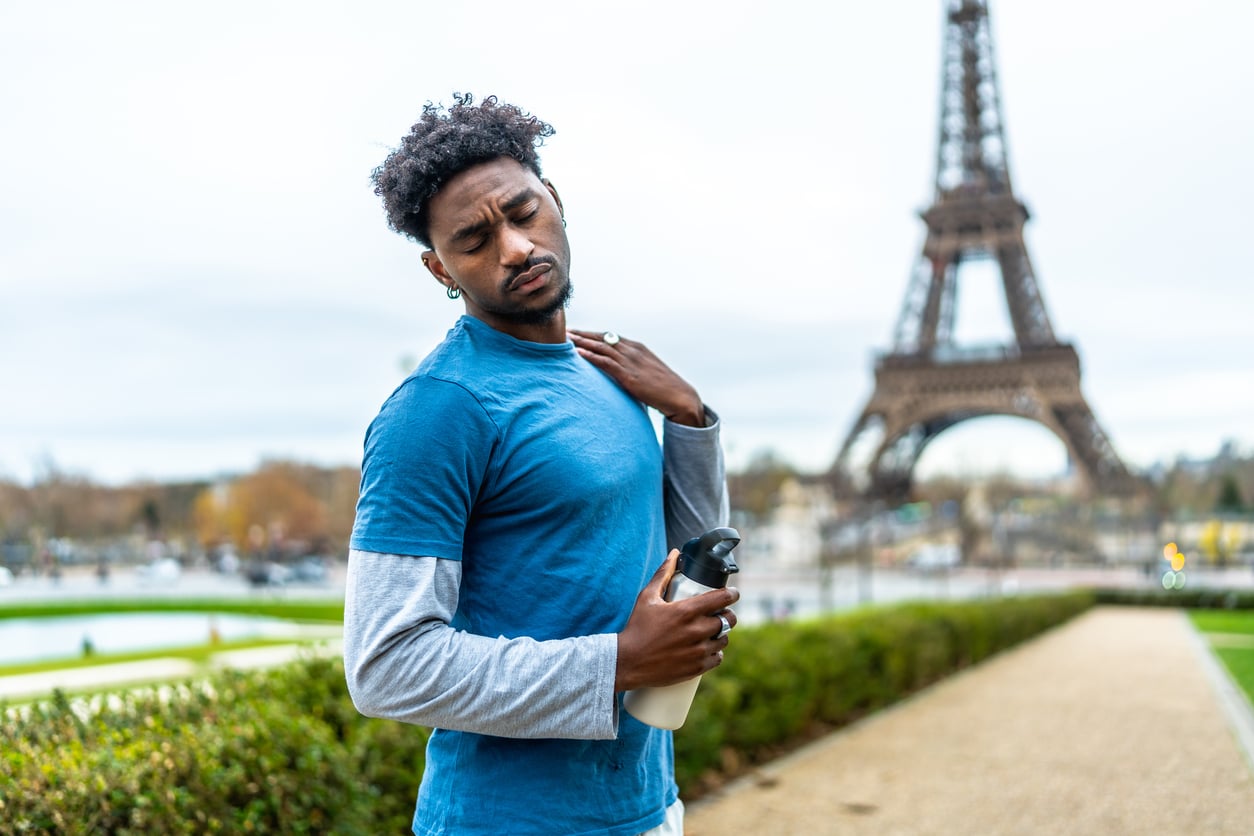 A man in athletic clothes holds his shoulder in discomfort while holding a water bottle, with the Eiffel Tower visible in the background.