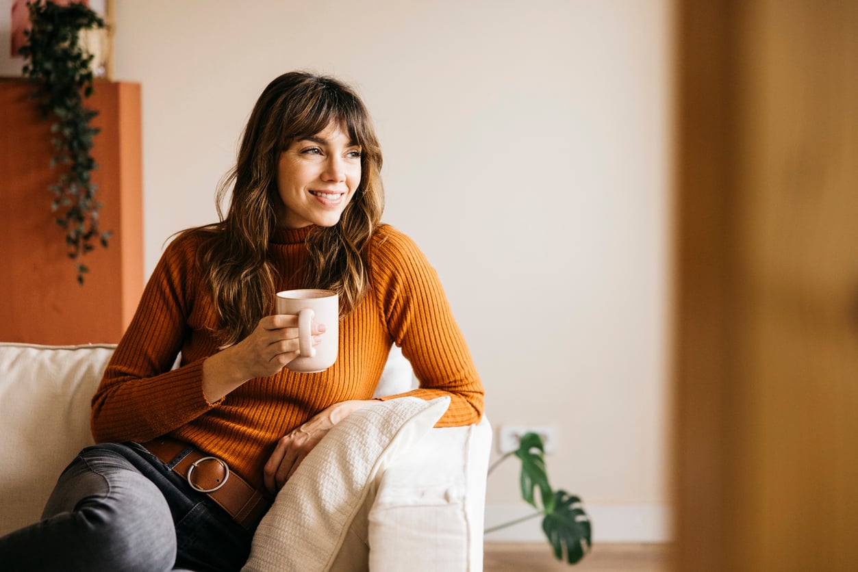A woman with brown hair, wearing an orange sweater and jeans, sits on a couch holding a mug and smiling, with a plant in the background.