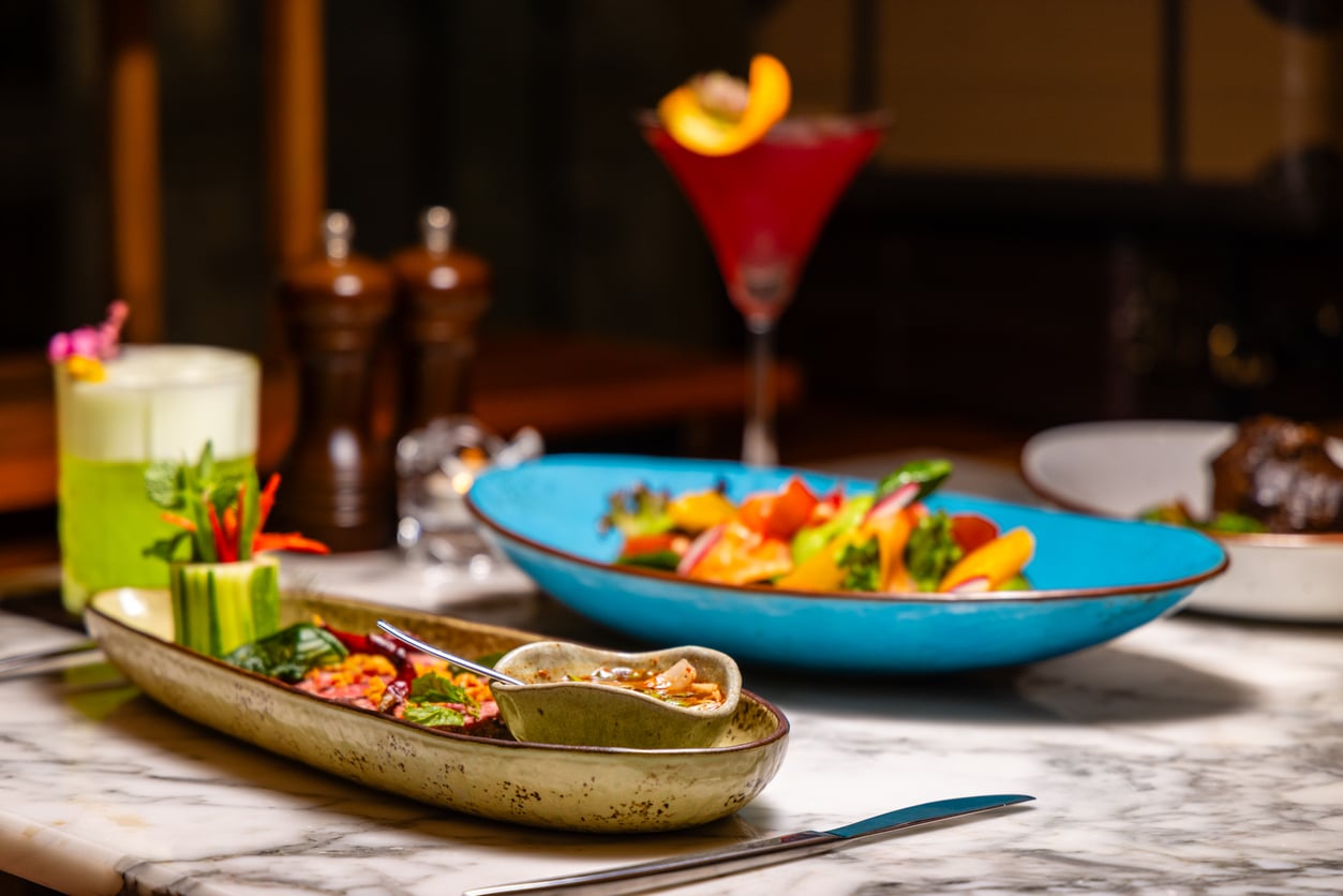 Colorful appetizers and a cocktail are arranged on a marble table, with fresh vegetables in a green dish, a salad in a blue bowl, and drinks in the background, creating a vibrant dining scene.