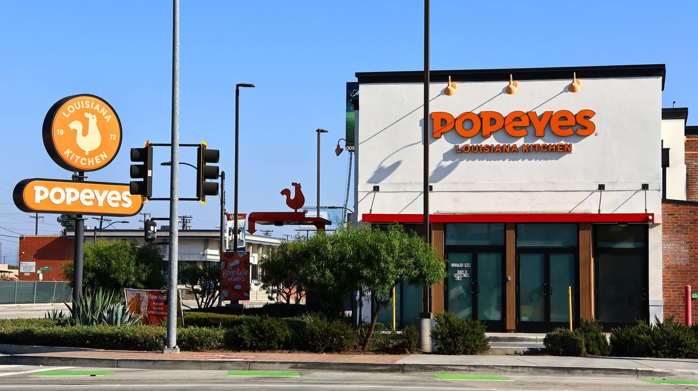 A Popeyes Louisiana Kitchen fast-food restaurant with orange signage, a chicken logo, large windows, and a drive-thru lane on a sunny day.