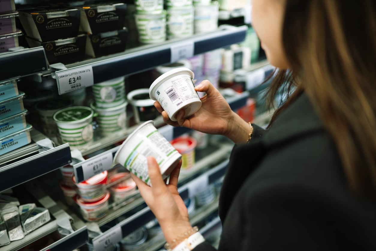 A person in a dark coat holds two containers, comparing labels while shopping for dairy products in a grocery store aisle—a scene that reflects mindful grocery habits. Shelves display various tubs and price tags, emphasizing the importance of making informed choices.