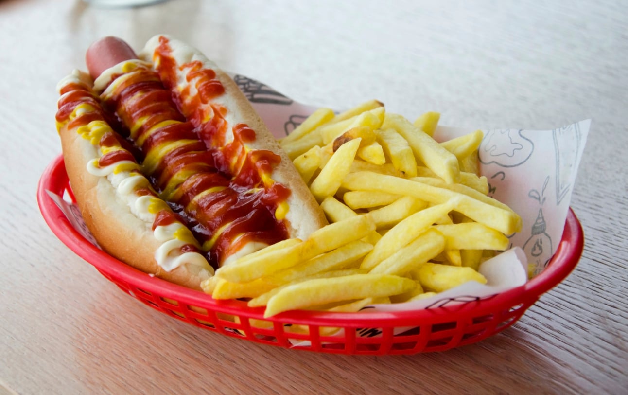 A red basket with a hot dog topped with ketchup, mustard, and mayonnaise, next to a serving of golden French fries on white parchment paper.