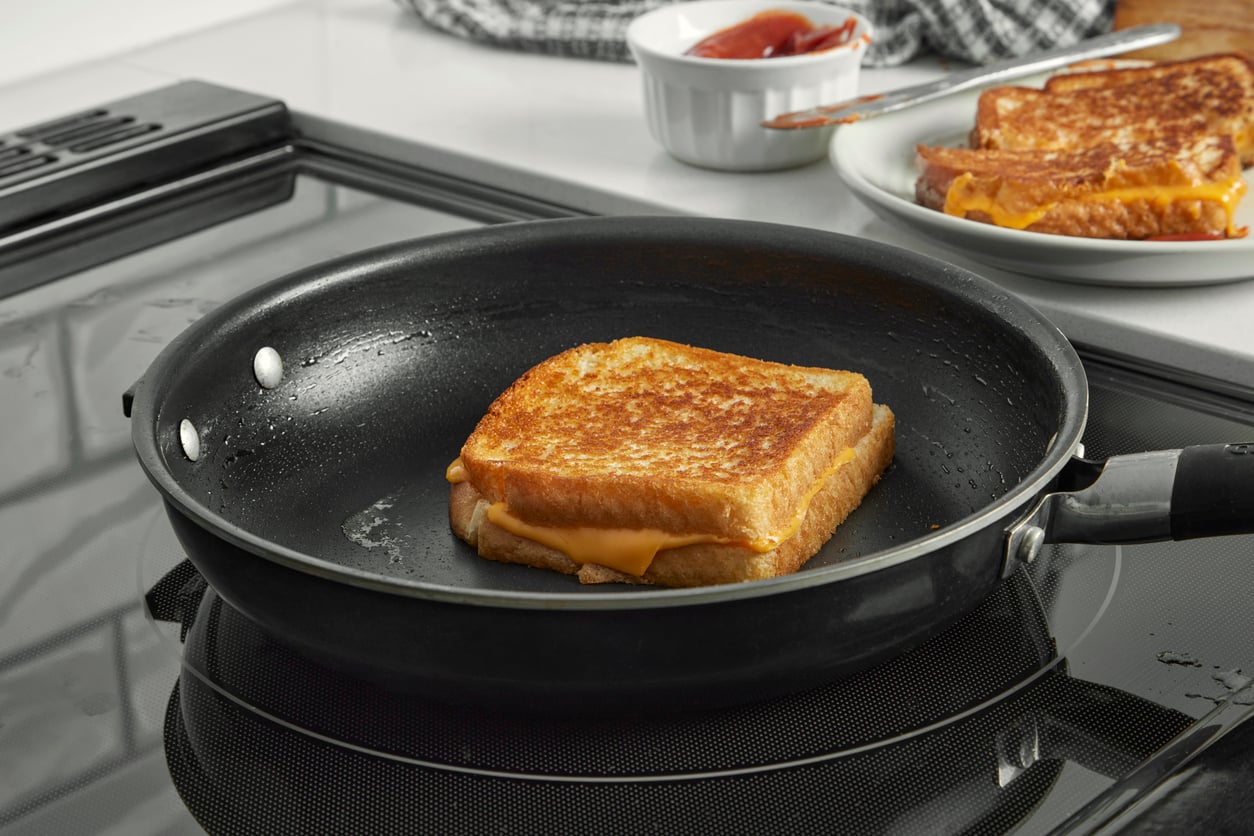 A grilled cheese sandwich cooking in a frying pan on a stovetop, with another sandwich and a small bowl of ketchup on a plate in the background.