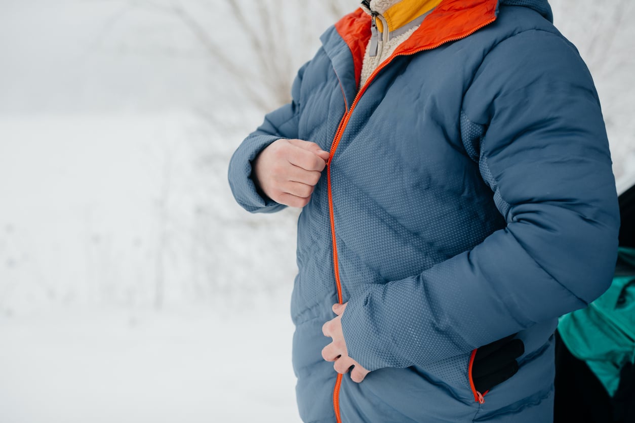 A person outdoors in snowy weather, wearing a blue puffer jacket with an orange lining, is zipping it up. Only the torso and hands are visible.