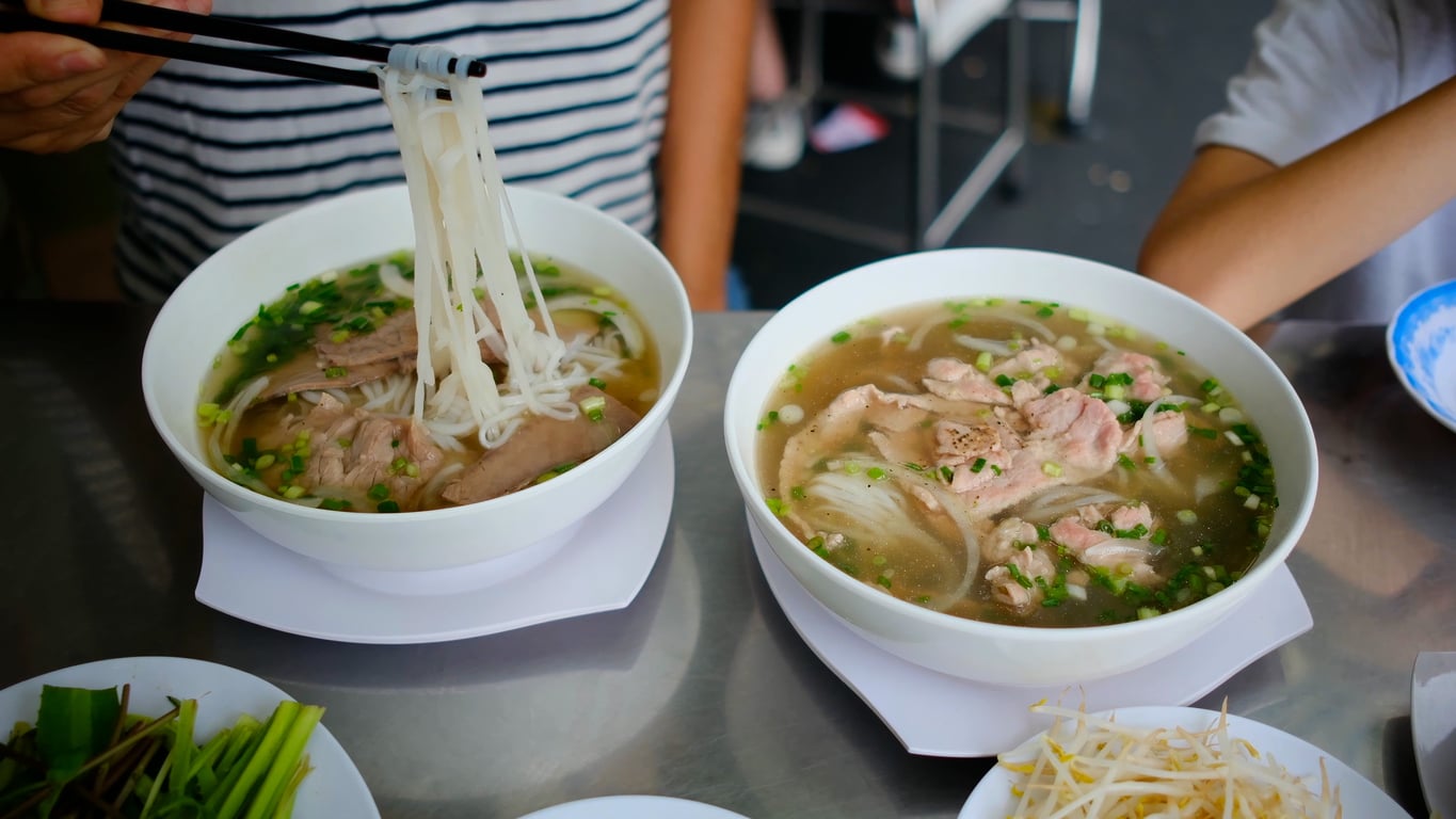 Two bowls of Vietnamese pho with noodles, sliced meat, and herbs. A person lifts noodles with chopsticks. Plates of fresh vegetables and bean sprouts are on the side.