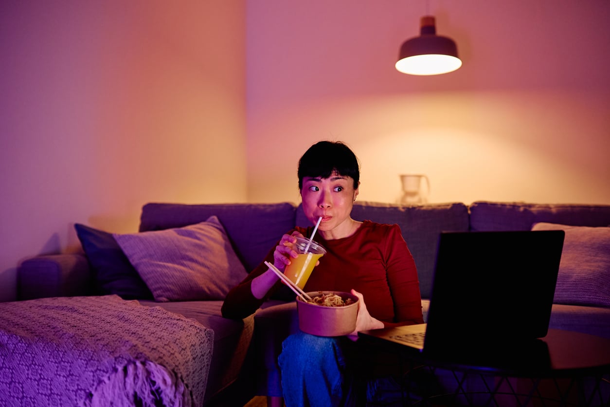 A person sits on a couch watching a laptop, holding a bowl of food and sipping a drink through a straw in a cozy, softly lit living room.