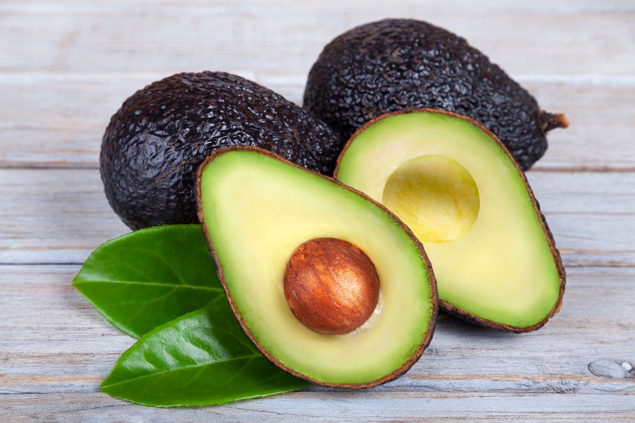 Two whole avocados and one avocado cut in half are displayed on a wooden surface with two green leaves, showing the seed and creamy green flesh inside—a beautiful example of how everyday foods that used to be considered fancy have become staples in our kitchens.