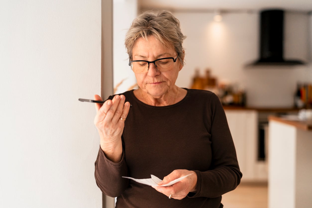 Older woman with short gray hair and glasses speaks into her smartphone while holding a piece of paper, standing in a modern kitchen with blurred appliances and counters in the background.