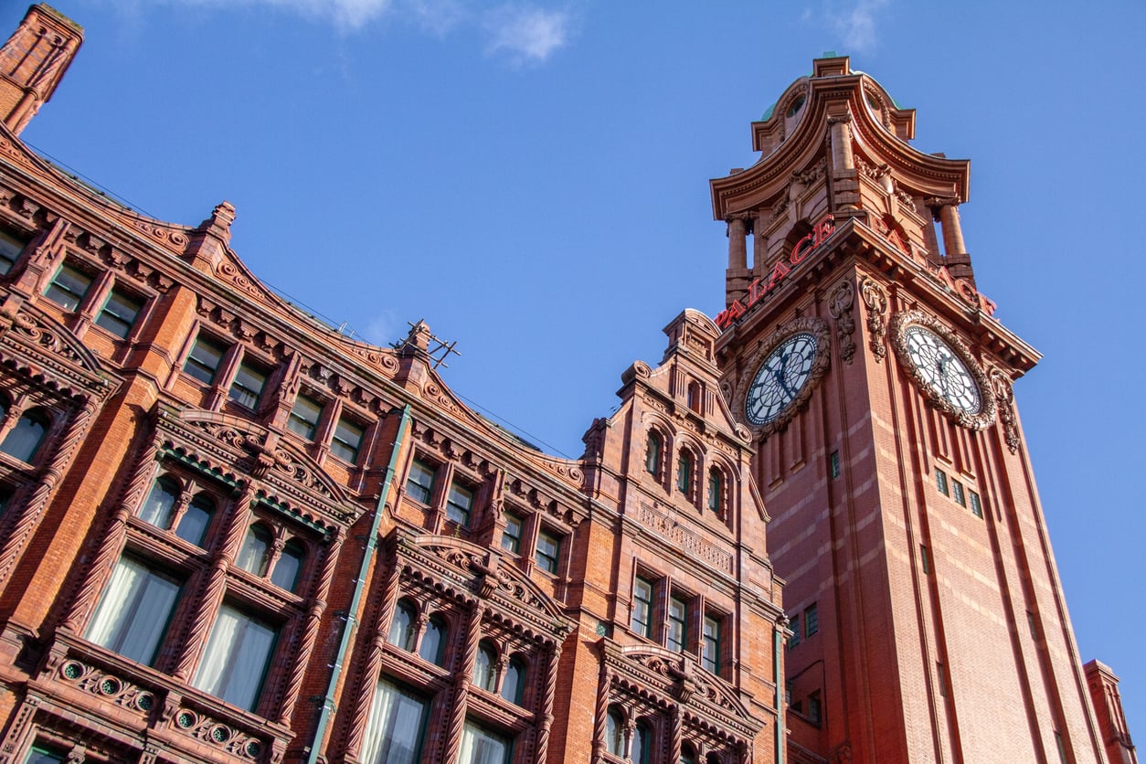 Red brick building with a tall clock tower against a blue sky. The structure features arched windows and detailed architectural elements, reminiscent of the elegance often found in distinguished hotel chains, with the word “REFUGE” displayed near the top of the tower.