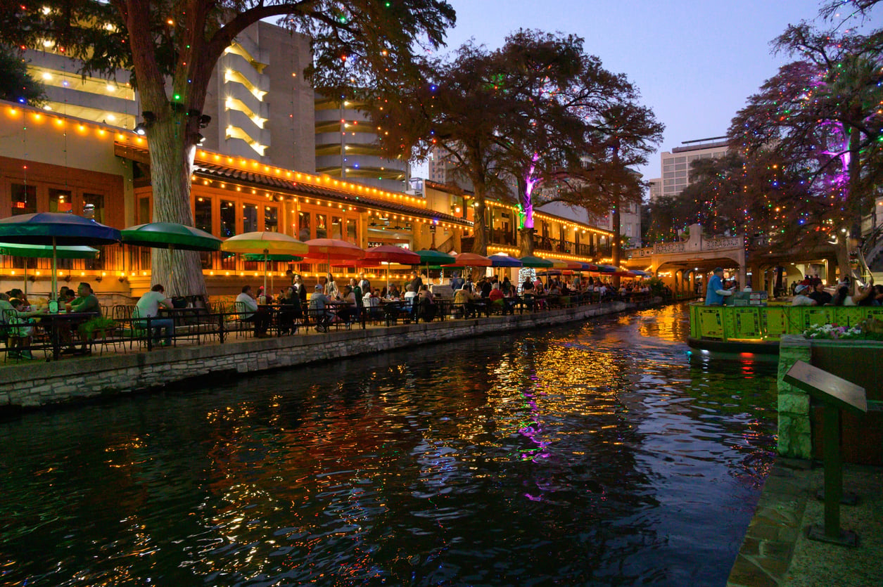 Colorful umbrellas shade diners along a lively riverside walkway at dusk, with festive lights in trees reflecting on the water, and boats passing by.