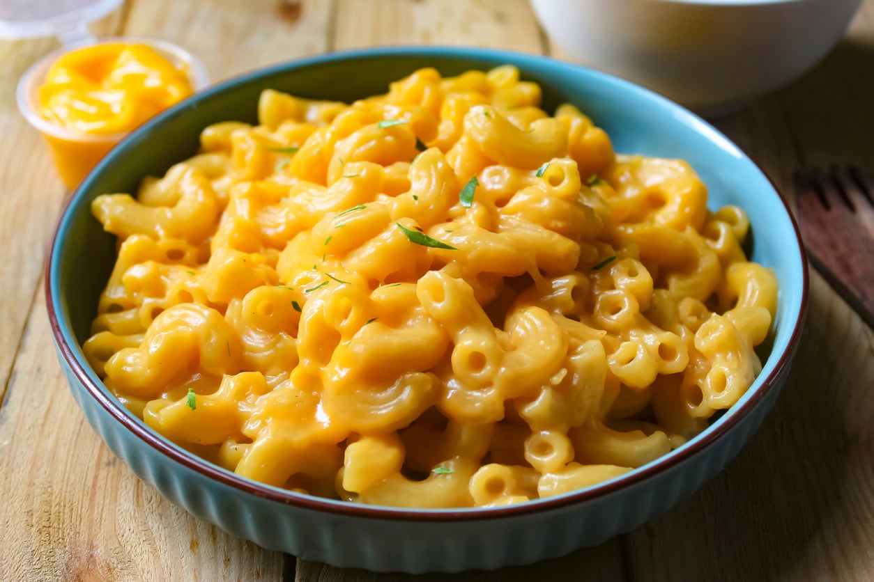 A bowl of creamy macaroni and cheese sits on a wooden table, garnished with a few green herbs. A small cup of extra cheese sauce and a white bowl are in the background.