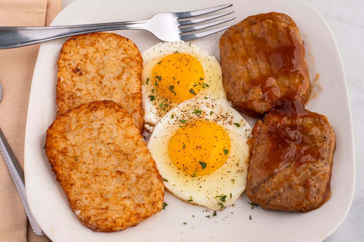 A plate with two hash brown patties, two sunny-side-up eggs topped with herbs, and two beef patties with brown gravy, plus a fork on the side.