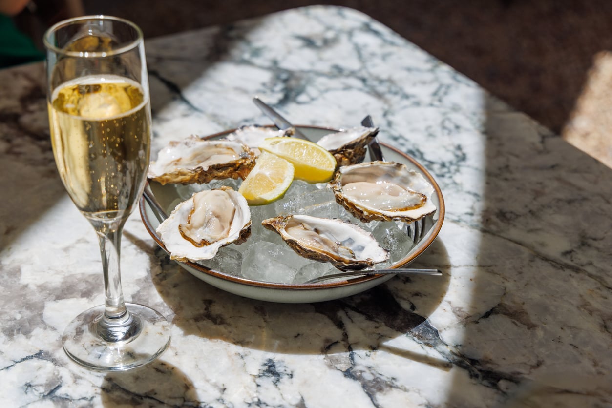 A glass of champagne sits next to a plate of oysters on ice with lemon wedges, placed on a marble table in sunlight—classic examples of foods people learn to appreciate later in life.