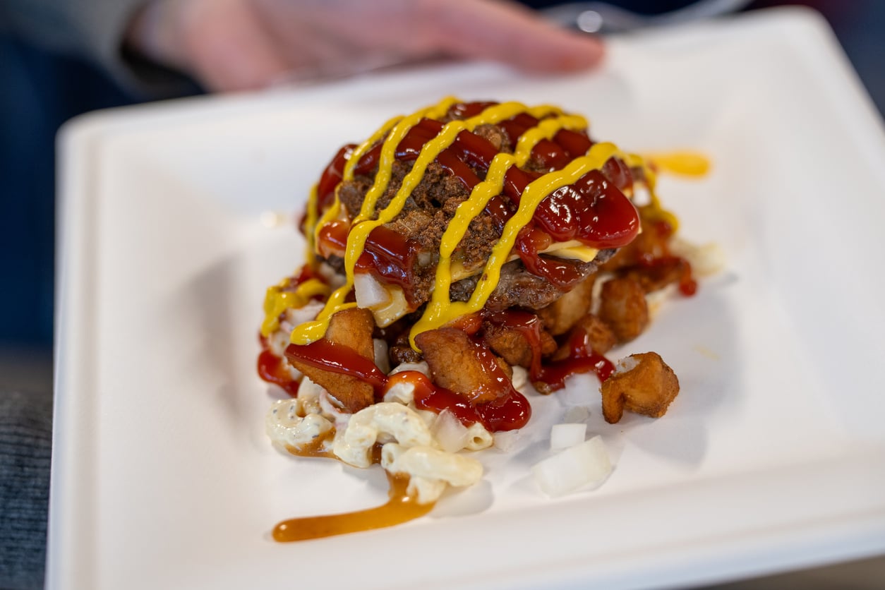 A plate of macaroni salad, home fries, chopped onions, and ground beef patties topped with melted cheese, ketchup, and mustard, served on a white tray.