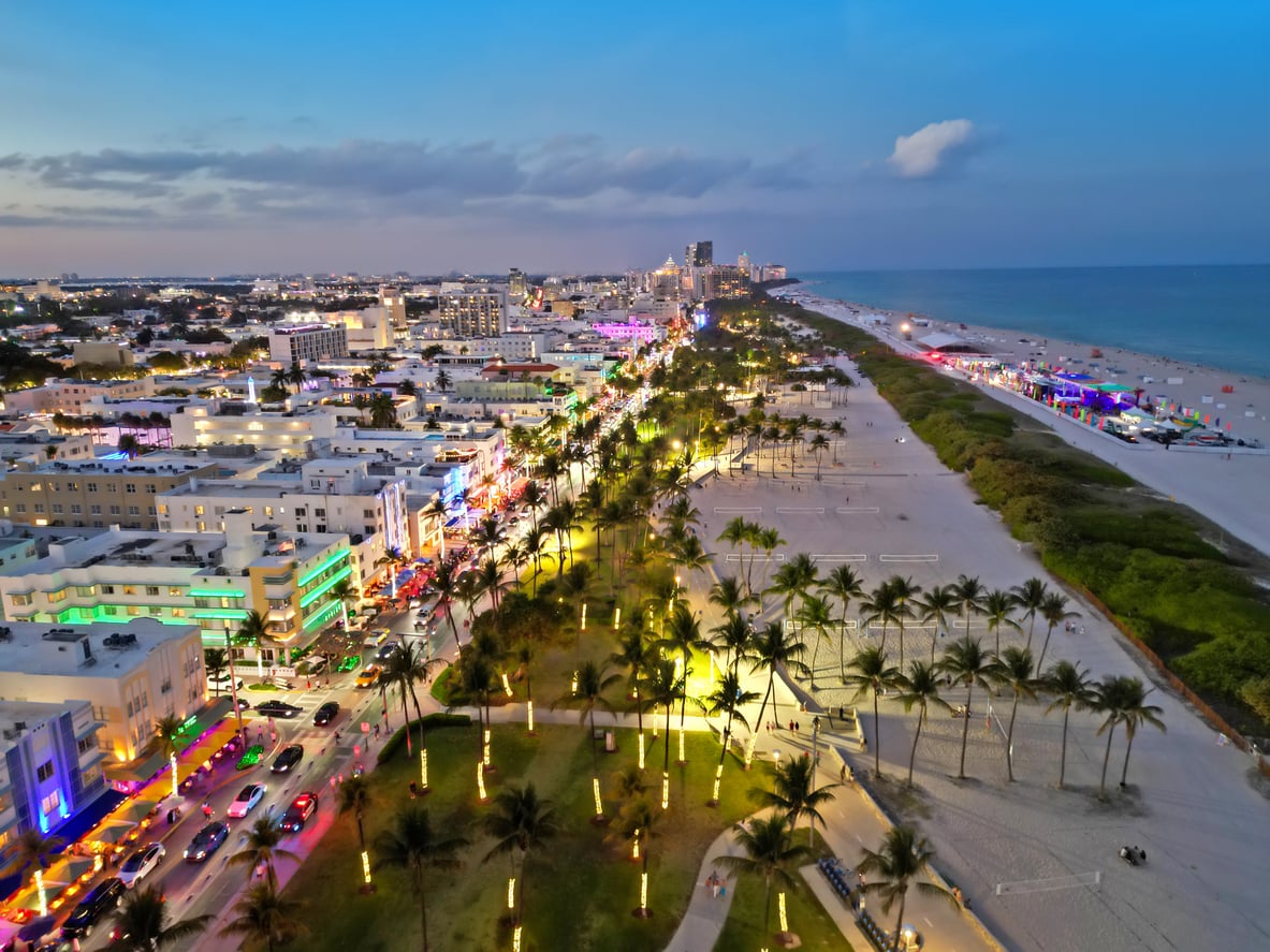 Aerial view of Ocean Drive in Miami Beach at dusk, with colorful lights, palm trees, busy streets, and sandy beach along the ocean.