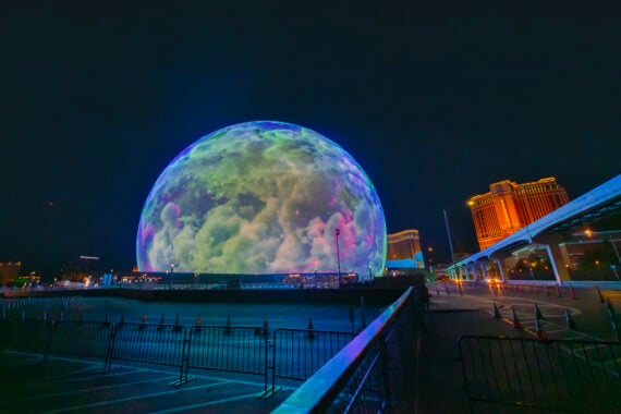 A large spherical building lit to resemble the moon at night, surrounded by metal barriers and city lights, with a modern hotel glowing orange in the background.