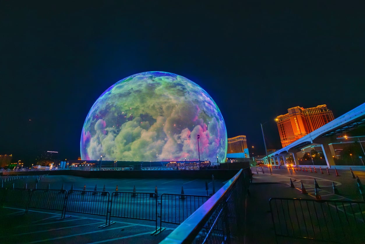 A large spherical building lit to resemble the moon at night, surrounded by metal barriers and city lights, with a modern hotel glowing orange in the background.