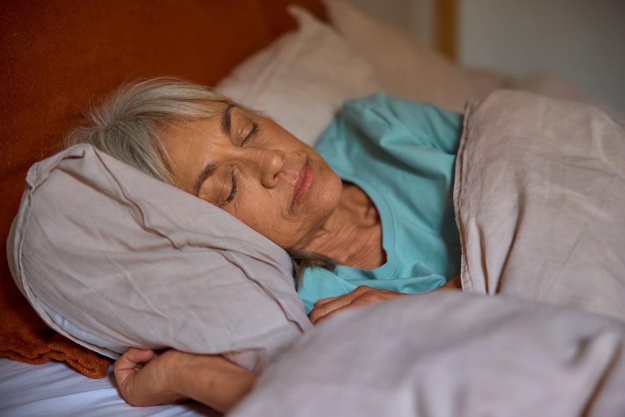 An older woman with gray hair sleeps peacefully on her side in bed, covered with a beige blanket and resting her head on a pillow.