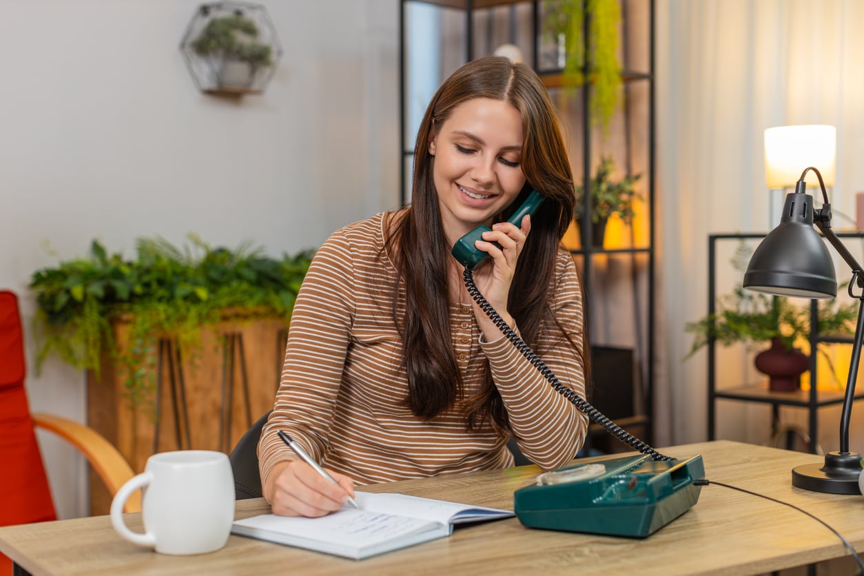 A woman with long brown hair smiles while talking on a green rotary phone and writing in a notebook at a desk with a white mug, lamp, and plants in the background.
