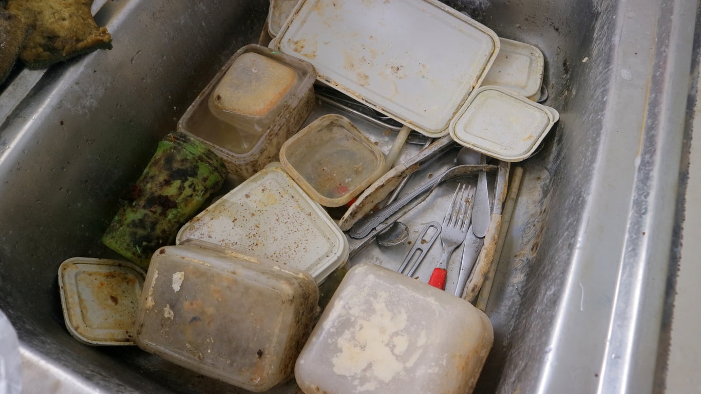 A kitchen sink filled with dirty plastic food containers, utensils, and cutlery, many of which have food residue and mold inside them.