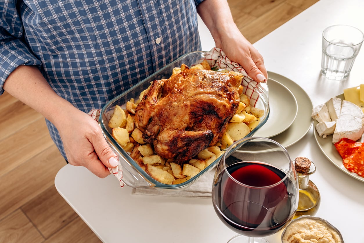 Person in a blue plaid shirt holding a glass dish with roasted chicken and potatoes; a table set with wine, cheese, and other food is visible.