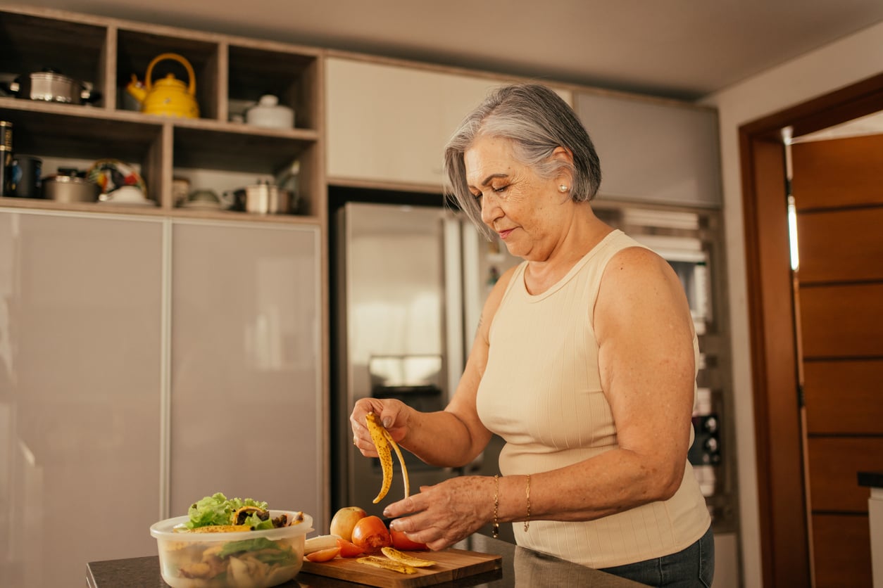 An older woman with gray hair stands in a kitchen, peeling a banana over a cutting board with fruits and vegetables. Shelves and a refrigerator are visible in the background.