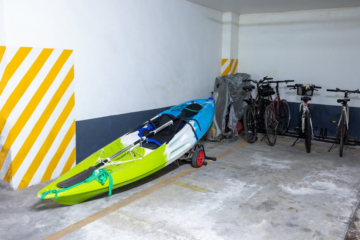A green and blue kayak with a paddle sits on a trolley in a parking garage, next to several bicycles, some covered with a gray tarp, against a wall with yellow striped markings.