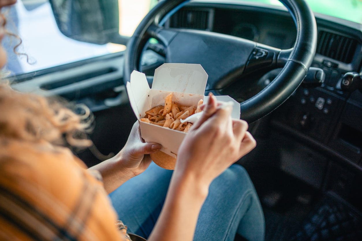 A person sitting in a vehicle’s driver seat eats pasta from a takeout container using a fork. The steering wheel and dashboard are visible in the background.