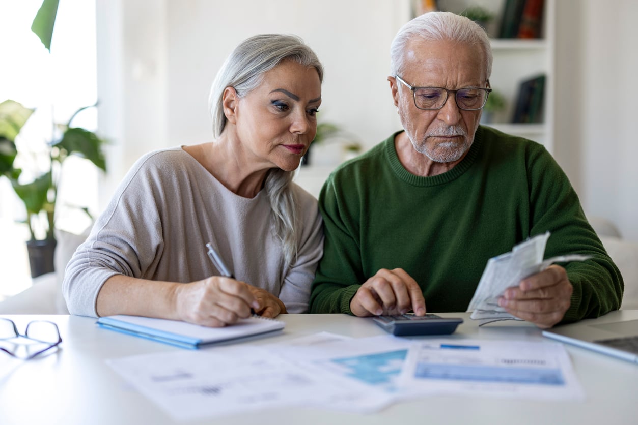 An elderly couple sits at a table, reviewing bills and using a calculator. The woman writes in a notebook while the man examines papers, both appearing focused and serious as they consider their retirement money habits. Financial documents are spread out on the table.