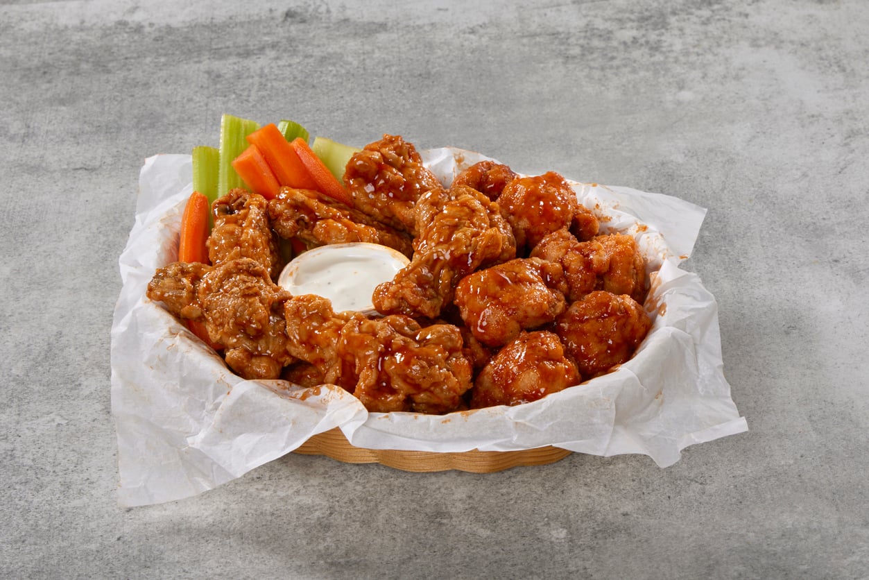 A basket of buffalo chicken wings with celery and carrot sticks, served with a cup of ranch dressing, on parchment paper and a gray stone surface.